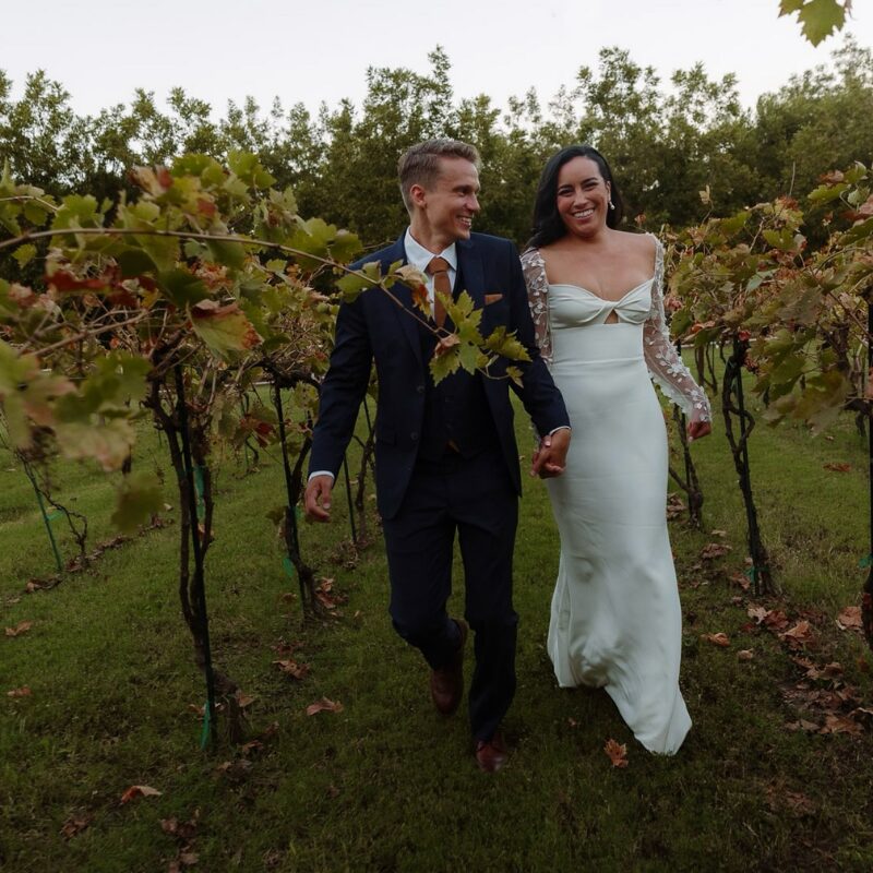 Bride and groom walk hand in hand through a vineyard, laughing together in the fall light.