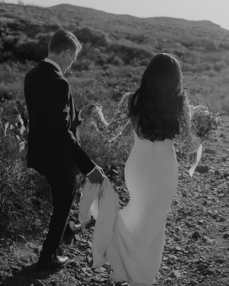 Black and white photo of a bride and groom walking hand in hand along a rocky desert trail.