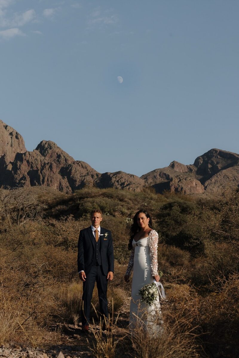 Bride and groom stand together in the desert brush as the moon rises above the Organ Mountains.