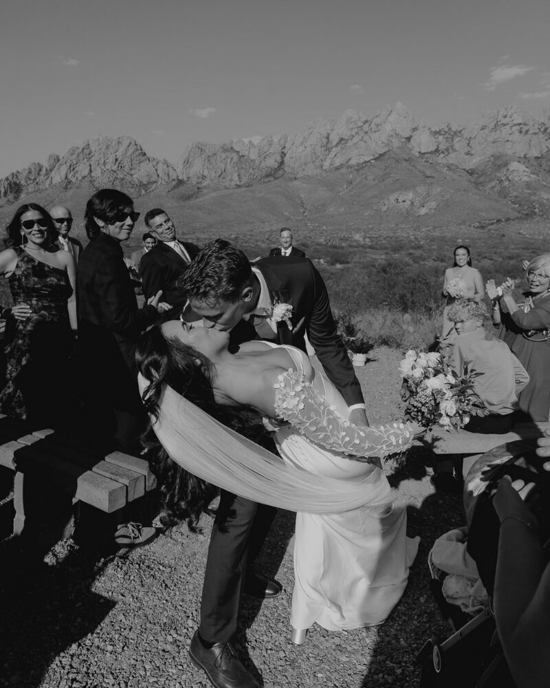 Black and white photo of a groom dipping his bride for a kiss during their ceremony, guests cheering, captured by their wedding day content creator.