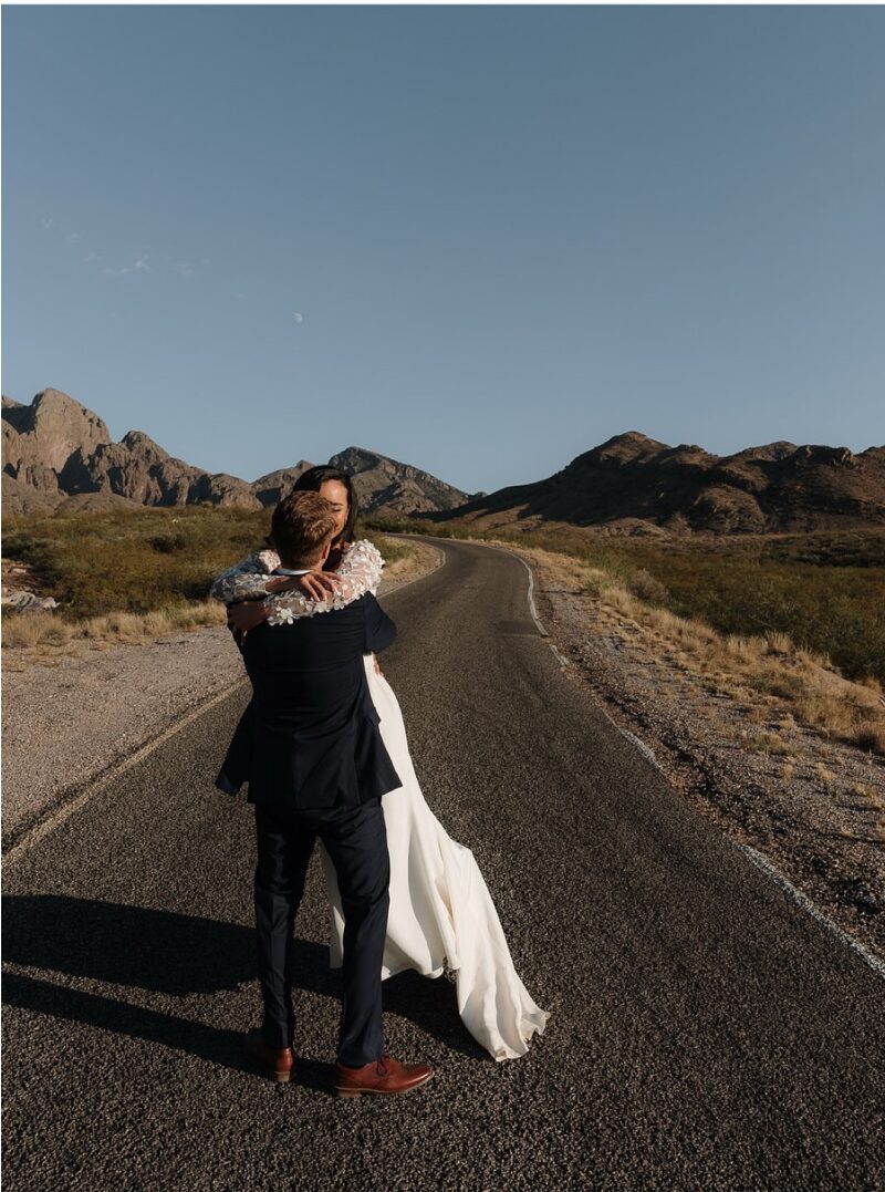 Groom embraces his bride on an open desert road as her dress train sweeps across the pavement.